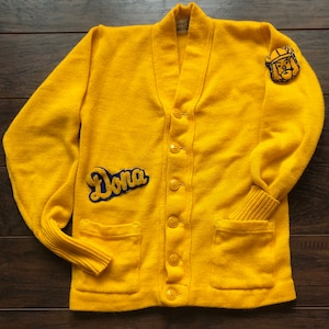 May include: A vintage yellow cardigan sweater with a V-neck, button closure, and two front pockets. The sweater features a "Dona" patch in blue and gold, along with a patch of a figure on the upper right sleeve. The sweater is made of a knitted material.