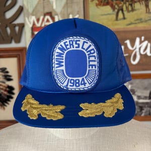 May include: A blue trucker hat with a mesh back and a curved brim. The front features a white and blue patch with the text "WINNER'S CIRCLE 1984". Gold embroidered leaf designs adorn the brim. A vintage-style cap.