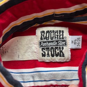 May include: Red and white striped shirt with a black and yellow stripe. The shirt has a label that says "Rough Stock" with the words "Panhandle Slim" underneath. The label also has a size tag that says "M 15 1/2 34/35".