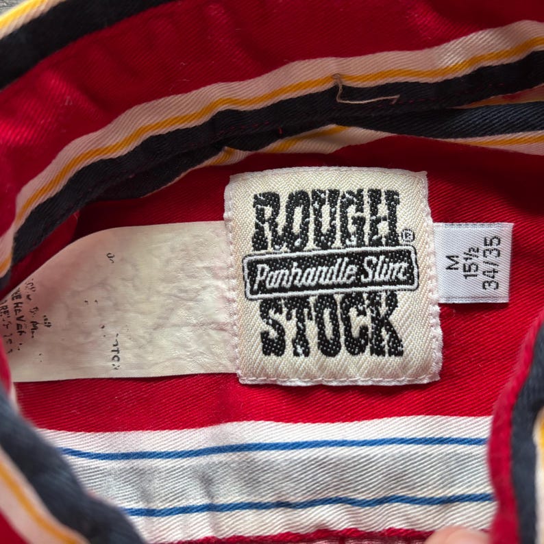 May include: Red and white striped shirt with a black and yellow stripe. The shirt has a label that says "Rough Stock" with the words "Panhandle Slim" underneath. The label also has a size tag that says "M 15 1/2 34/35".