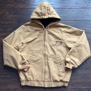 May include: A tan hooded jacket with a black interior lining. The jacket has a zip closure, two front pockets, and ribbed cuffs and hem. The jacket features an embroidered design on the chest.