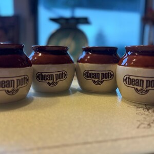 Peut inclure: Quatre petits pots à haricots en céramique brun et blanc avec les mots "Bean Pot" imprimés sur le devant.