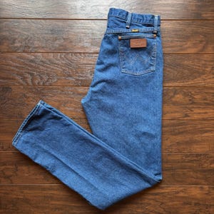May include: Blue denim jeans with a classic five-pocket design. The jeans feature a brown leather patch on the back pocket with the brand name. The jeans are laid flat on a wooden surface.