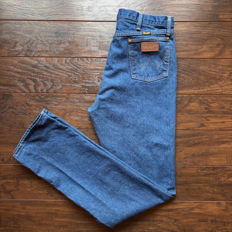 May include: Blue denim jeans with a classic five-pocket design. The jeans feature a brown leather patch on the back pocket with the brand name. The jeans are laid flat on a wooden surface.