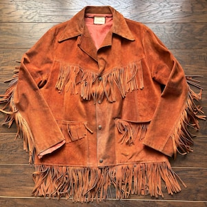 May include: A rust-colored suede jacket with long fringe detailing. The jacket features a collared neckline, button closures, and pockets. The fringe is along the sleeves, chest, and hemline. The jacket is laid flat on a wooden surface.