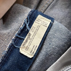 May include: Close-up of a pair of blue denim Wrangler jeans. The jeans' label is visible, displaying the brand name, size, and care instructions. The label reads "Made in USA" and "100% Cotton."