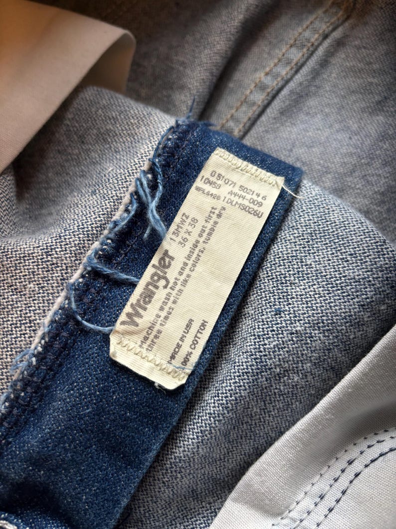 May include: Close-up of a pair of blue denim Wrangler jeans. The jeans' label is visible, displaying the brand name, size, and care instructions. The label reads "Made in USA" and "100% Cotton."