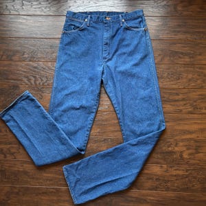 May include: Blue denim jeans with a classic five-pocket design. The jeans have a button and zipper closure, and are made of a durable denim fabric. The jeans are laid flat on a wooden surface.