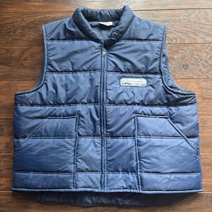 May include: A navy blue quilted vest with a full-length zip and two front pockets. The vest features a small patch with text and a graphic. The vest is made of a smooth, shiny material.
