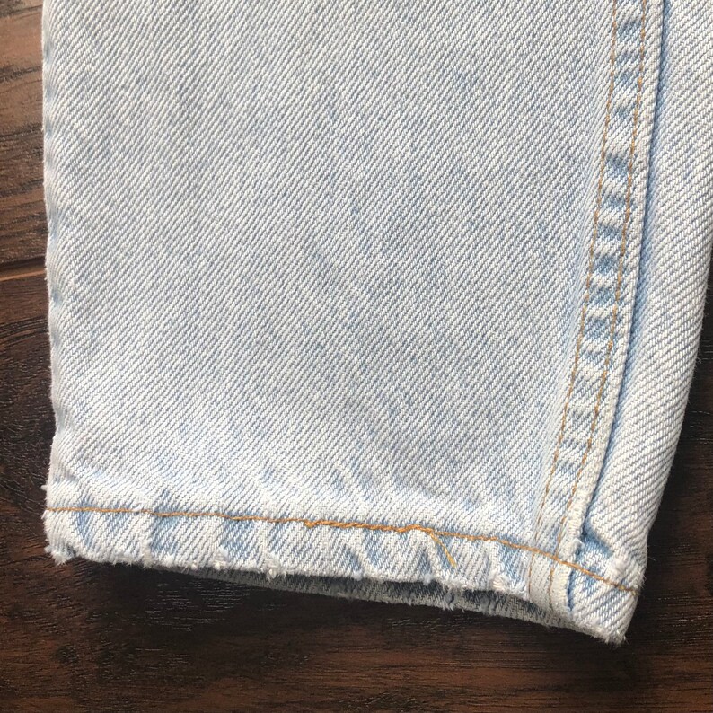 May include: Close-up of the hem of light blue denim jeans with a raw edge and a single stitch of light brown thread.