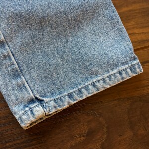 May include: Close-up of the hem of a pair of light blue denim jeans. The fabric is a light wash with a slightly distressed look. The stitching is visible along the edge of the hem.