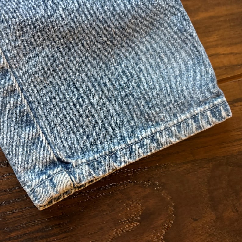 May include: Close-up of the hem of a pair of light blue denim jeans. The fabric is a light wash with a slightly distressed look. The stitching is visible along the edge of the hem.