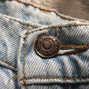 May include: Close-up of a light blue denim jean with a copper-coloured button that reads "LEVI STRAUSS & CO. SAN FRANCISCO. CAL."