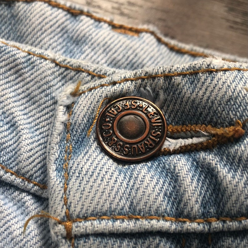 May include: Close-up of a light blue denim jean with a copper-coloured button that reads "LEVI STRAUSS & CO. SAN FRANCISCO. CAL."