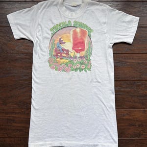 May include: White t-shirt featuring a graphic of a cartoon cowboy riding a donkey in front of a sunset with the text "Tequila Sunrise".