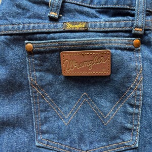May include: Close-up of the back pocket of blue denim jeans. The pocket has a stitched, stylized 'W' design. A brown leather patch with the brand name 'Wrangler' is centered on the pocket. The jeans have a classic, vintage style.