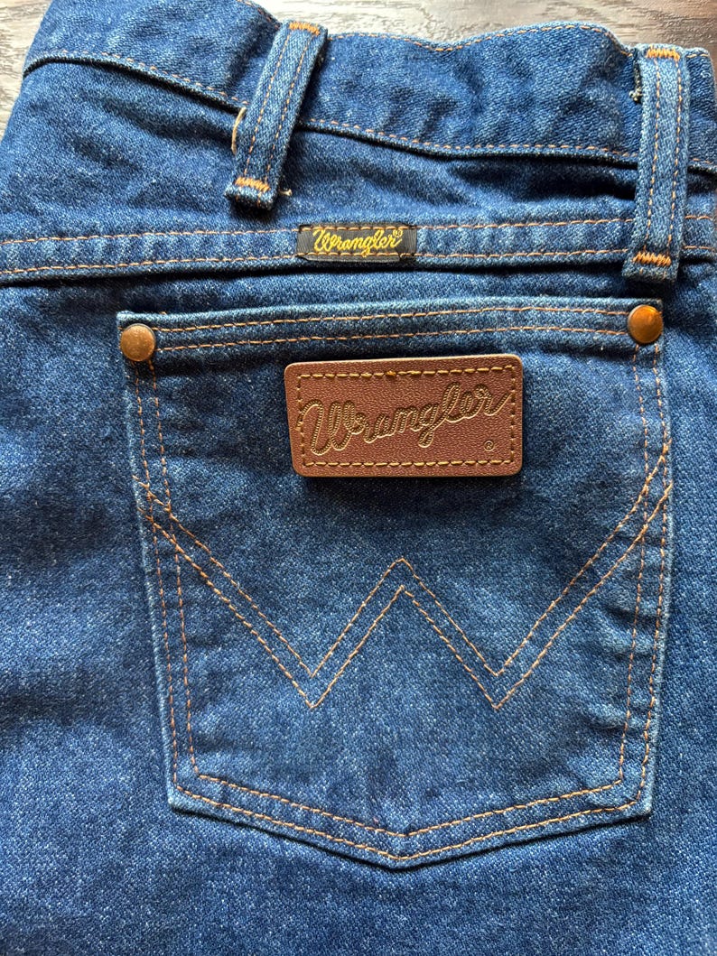 May include: Close-up of the back pocket of blue denim jeans. The pocket has a stitched, stylized 'W' design. A brown leather patch with the brand name 'Wrangler' is centered on the pocket. The jeans have a classic, vintage style.