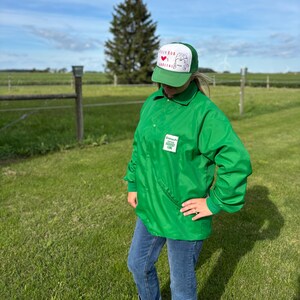May include: A green nylon jacket and a green and white trucker hat. The hat features the text "Billy Bob" and a heart graphic. The jacket has a patch that reads "Beach Grove Harvest Club". The person is wearing blue jeans.