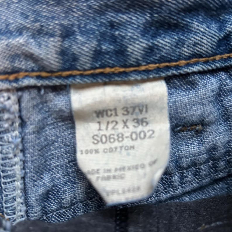 May include: A close-up of a blue denim label with white text that reads "WC1 37VI 1/2 X 36 S068-002 100% COTTON MADE IN MEXICO OF FABRIC".