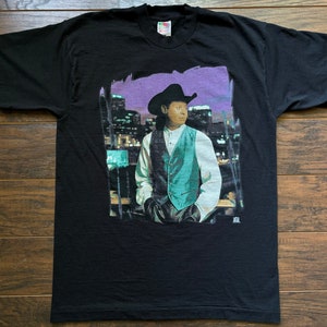 May include: Black t-shirt with a graphic print of a man wearing a cowboy hat and vest, standing in front of a city skyline at night.