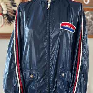 May include: Navy blue vintage jacket with a zipper closure, two front pockets, and a stand-up collar. The jacket features red and white stripes down the sleeves and a "PUROLATOR" patch on the chest. A classic retro style.