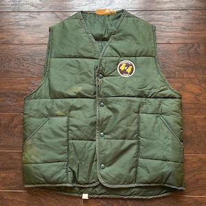 May include: An olive green quilted vest with a button-up front and two front pockets. A circular patch with the text "4x4 FEEDS" is on the upper right chest. The vest is made of a durable, possibly water-resistant material.