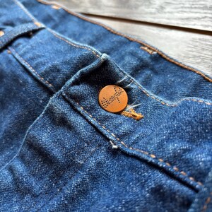 May include: Close-up of blue denim fabric with a brown button. The button is round and has a detailed design. The denim has visible stitching in a contrasting orange color.