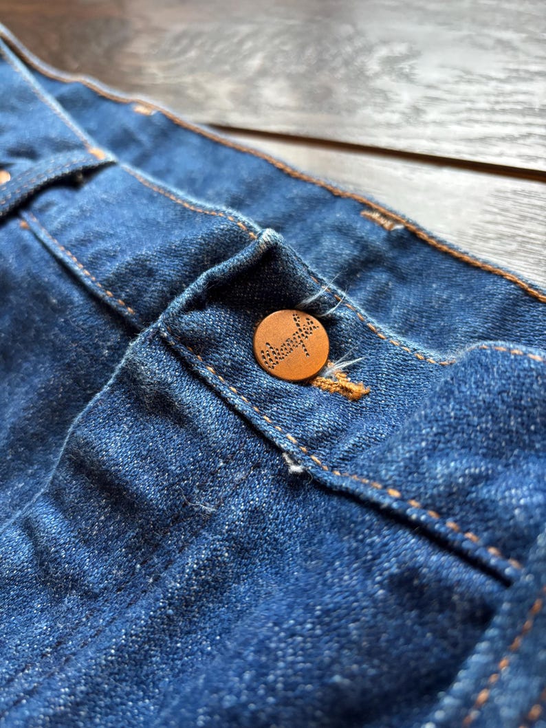 May include: Close-up of blue denim fabric with a brown button. The button is round and has a detailed design. The denim has visible stitching in a contrasting orange color.