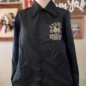 May include: Black nylon jacket with a button-up front, collared neckline, and two front pockets. The jacket features white text and graphics on the chest that read "ATOMIC Worlds Fastest SPEEDWAY Knoxville, TN."