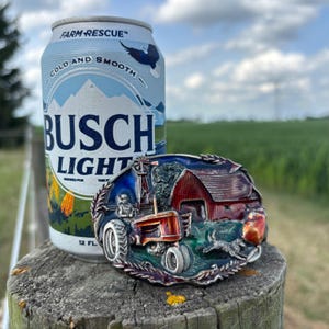 May include: A can of Busch Light beer and a detailed metal belt buckle. The buckle depicts a farm scene with a red barn, tractor, and running dog. The beer can is light blue with the words "Busch Light" in large letters.