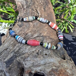 Puede incluir: Un brazalete colorido hecho de cuentas de papel enrollado con cuentas negras intercaladas. El brazalete está sobre un trozo de madera a la deriva.