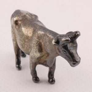 May include: A small, silver-toned cow figurine. The miniature bovine has a detailed form, with a slightly weathered appearance. The cow is standing, with its head turned slightly to the right. The figurine is likely a decorative item or collectible.