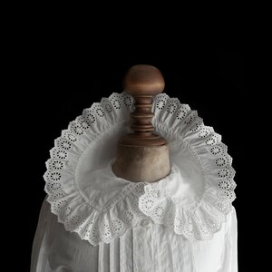 Puede incluir: Una blusa de algodón blanco con un gran cuello de encaje. El cuello está hecho de encaje blanco delicado con un borde festoneado. La blusa es un estilo clásico con un diseño simple.