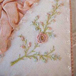 May include: A close-up of a white fabric with delicate pink and blue embroidered flowers. The fabric is trimmed with a row of small, clear beads.