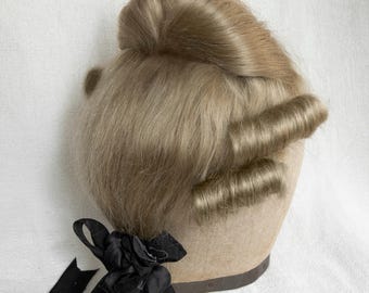 Könnte beinhalten: Eine blonde Perücke mit einem Dutt oben und zwei großen Locken an den Seiten. Die Perücke sitzt auf einem Mannequin-Kopf und hat ein schwarzes Band, das um die Basis gebunden ist.
