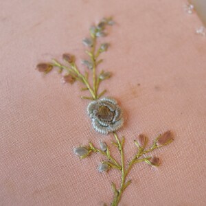 May include: A delicate floral embroidery design on a pale pink fabric. The design features a small flower with a light blue centre and green stems with small pink buds.