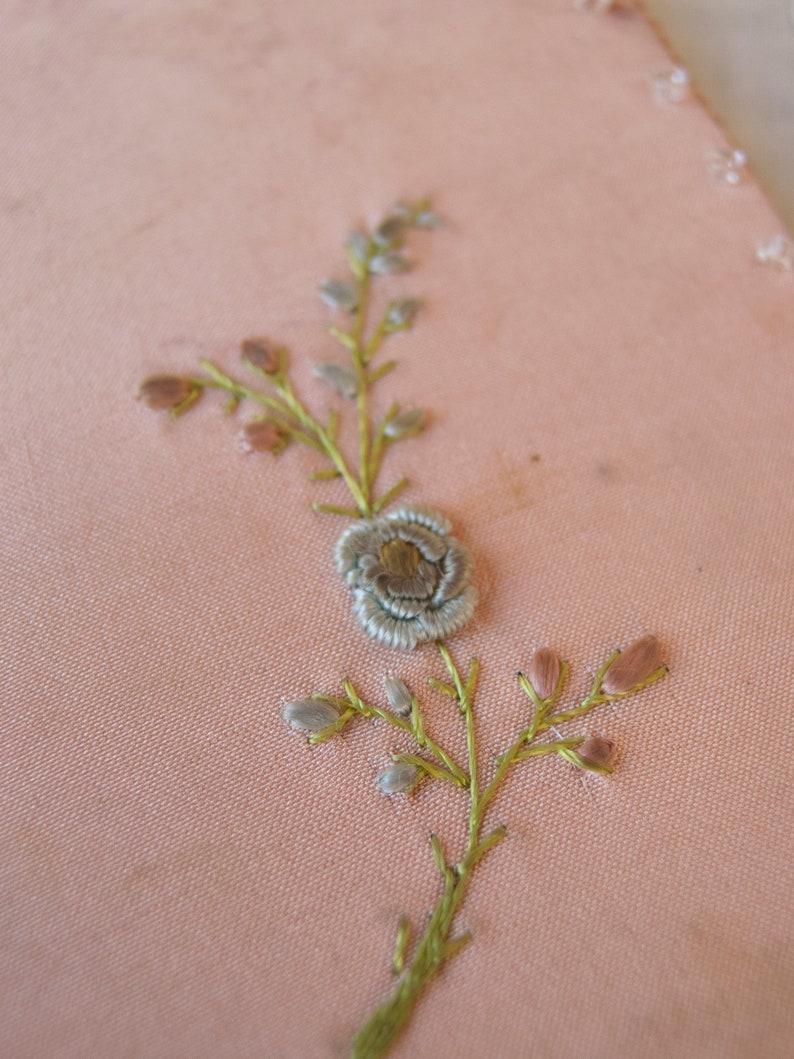 May include: A delicate floral embroidery design on a pale pink fabric. The design features a small flower with a light blue centre and green stems with small pink buds.