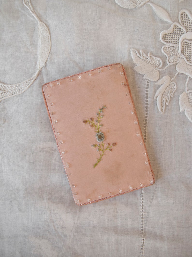 May include: A pink rectangular box with a beaded border and a delicate floral design embroidered on the front.