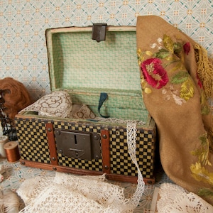 Peut inclure: Un coffre ancien ouvert avec un motif à carreaux noirs et jaunes, rempli de dentelle et de tissu. Un châle marron avec broderie florale et franges est drapé sur le côté. Des bobines de fil et une perruque sont également visibles.
