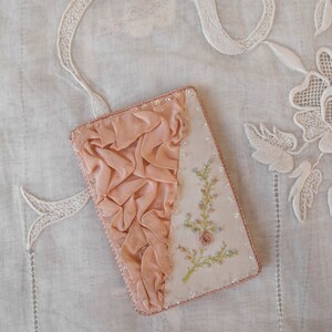 May include: A small pink fabric covered box with a delicate floral embroidery design. The box is decorated with a ruffled fabric trim and has a beaded border.