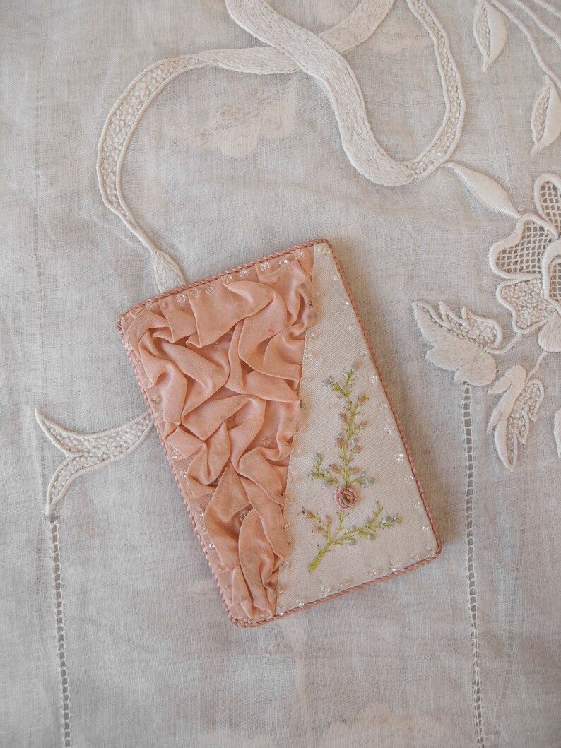 May include: A small pink fabric covered box with a delicate floral embroidery design. The box is decorated with a ruffled fabric trim and has a beaded border.