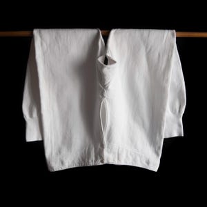 Op de afbeelding: Een wit shirt met lange mouwen hangt aan een houten roede tegen een zwarte achtergrond. Het shirt is dichtgeknoopt en heeft een klassiek ontwerp. De mouwen hebben manchetten.