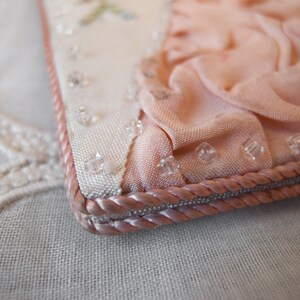 May include: A close-up of a pink fabric purse with a white fabric lining and a pink rope trim. The purse is decorated with clear beads.