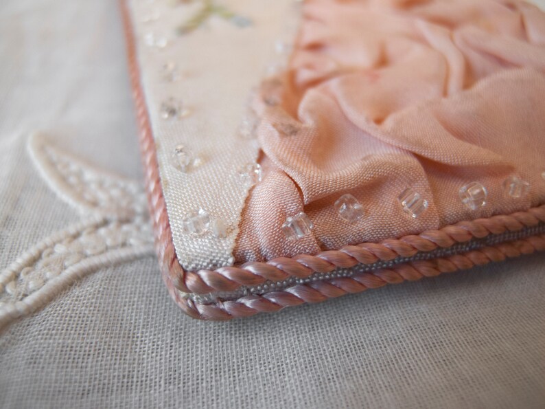 May include: A close-up of a pink fabric purse with a white fabric lining and a pink rope trim. The purse is decorated with clear beads.