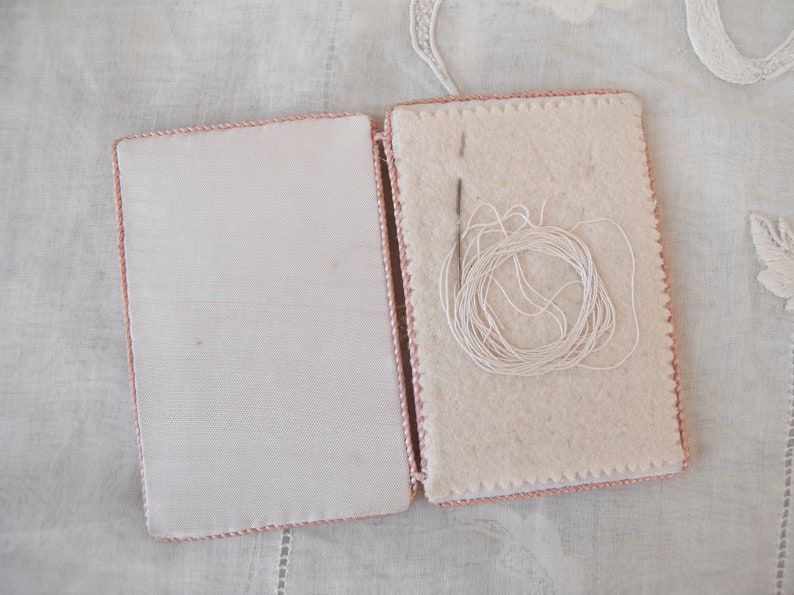 May include: A white needle and thread are inside a pink fabric needle case with a white background.