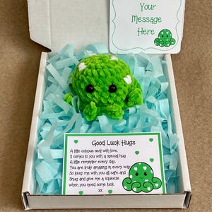May include: A green crocheted octopus with white spots is presented in a box with blue paper. The box contains a card that reads "Good Luck Hugs" and another card that says "Your Message Here".
