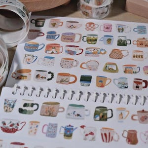 Pode incluir: Uma folha de autocolantes com ilustrações de chávenas de café e canecas em várias cores e padrões. Rolos de fita washi a condizer são visíveis no fundo, juntamente com um calendário de blocos de madeira.