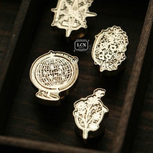 May include: Four brass stamp designs in a wooden drawer. The designs include a compass, a globe, and two floral designs.
