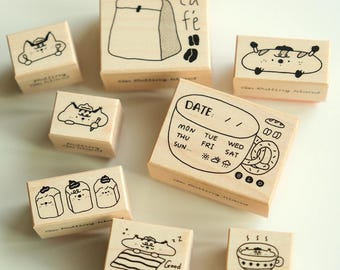 May include: A collection of wooden stamps with various black ink illustrations. Designs include a coffee bag, a date stamp, and several cute character designs. The text "Cafe" and "Good Night" are visible on some stamps.