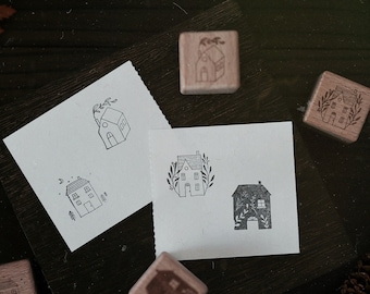 May include: Five wooden stamps with different house designs. The stamps are on a dark brown surface with a piece of paper with a house design on it.